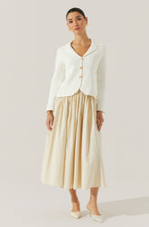 A woman is wearing a cream-colored, long-sleeved blazer with a notched collar and four large buttons down the front. The blazer has a slightly cropped fit with a pointed hem. She pairs it with a high-waisted, full-length, beige pleated skirt that reaches her ankles. She is also wearing cream-colored pointed-toe heels.